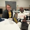 Dr. Elyse Stevens wears glasses, a yellow t-shirt and a sweater. Ronald Major wears a stylish white track suit.