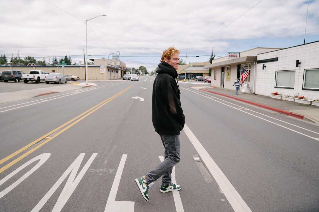 Cody Coughenhour crosses a street in Port Angeles, Wash. He's wearing a black hoodie and a cloudy sky is visible in the distance.