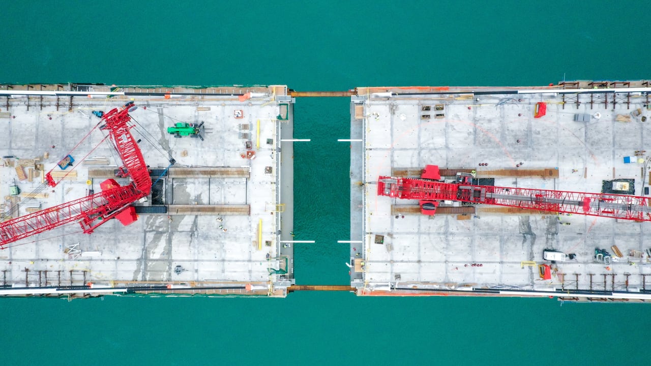 An aerial shot of a bridge under construction. there is a gap in the middle but the two sides are connected at the edges by beams.