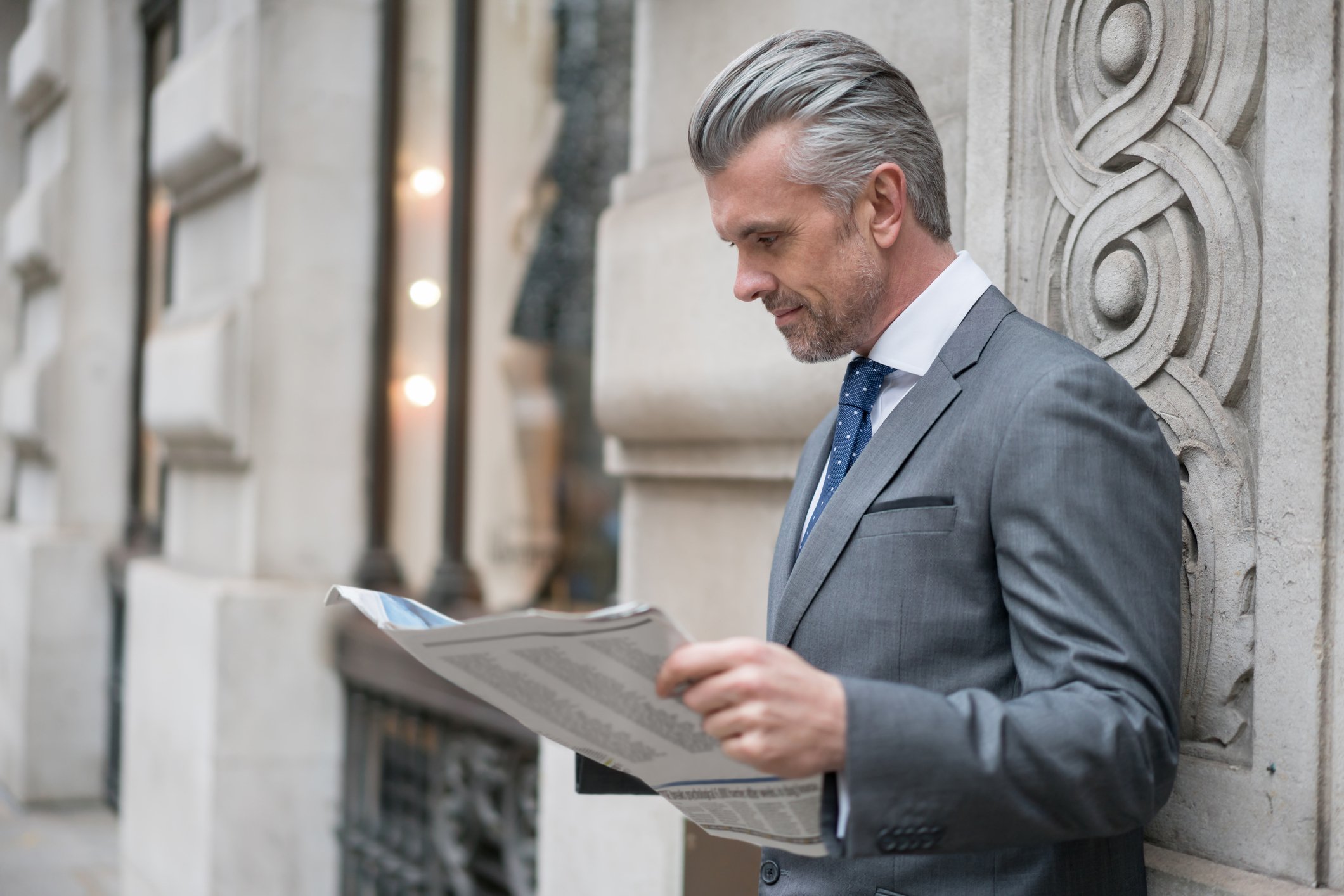 A person in a gray suit reads a newspaper while leaning against a stone wall.