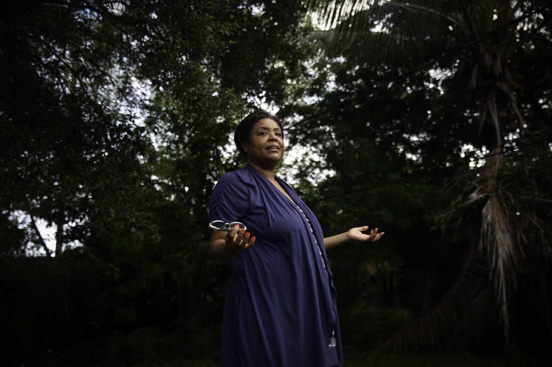 Wearing a dark blue dress, Farah Larrieux stands in her backyard in South Florida. Multiple trees stand in the background.