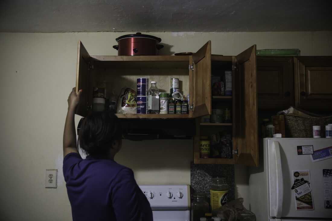 Photographed with her back to the camera, Farah Larrieux opens a kitchen cabinet that's above a stove. The cabinet is filled with pantry staples.