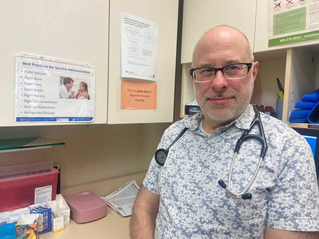 Pediatrician John Snyder is seated at a pediatrics practice. A stethoscope is slung over his shoulders. Wall cabinets are in the background.
