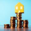 This photo shows four stacks of coins of varying heights standing in a row. At the top of the tallest stack, the second stack from the right, sits a small model of a yellow house with a front door and two windows. The background of the photo is light blue.