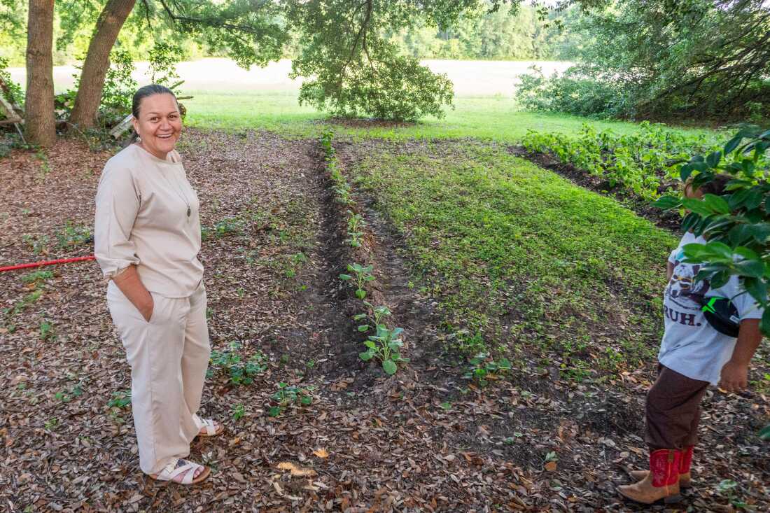 Angie Lowery is shown smiling, standing at the edge of a small vegetable garden, with one row of small green plants visible in several furrowed rows of earth.
