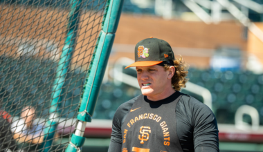 A baseball player in a black San Francisco Giants shirt and cap stands near a green batting cage on a sunny day.