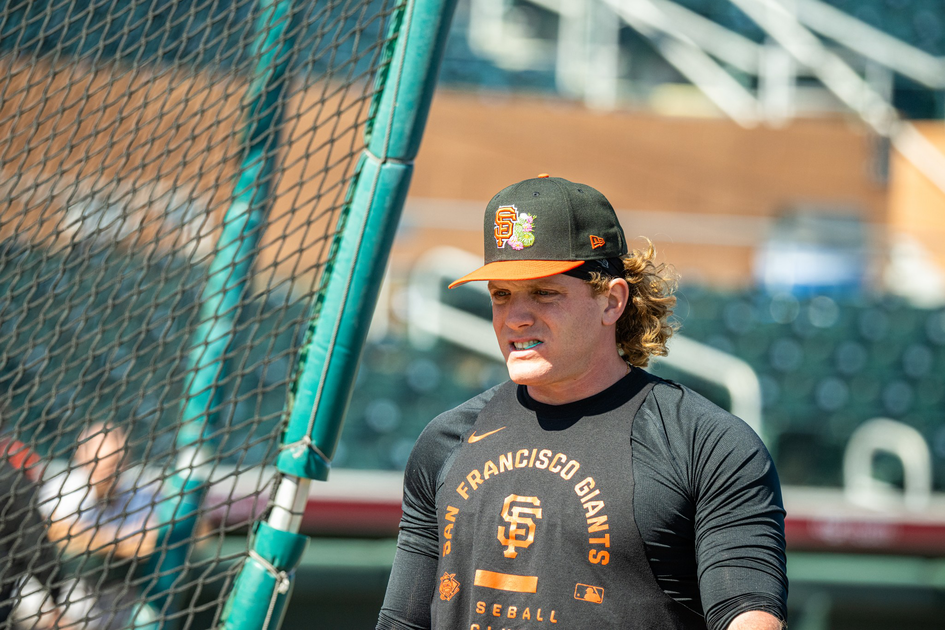 A baseball player in a black San Francisco Giants shirt and cap stands near a green batting cage on a sunny day.