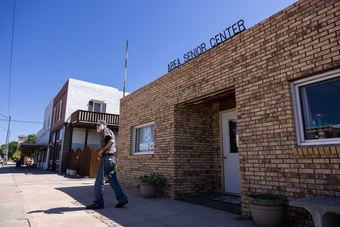 A man with a white beard is shown leaving a one-story brick building on a small town street. The building has a sign that reads, "Area Senior Center."