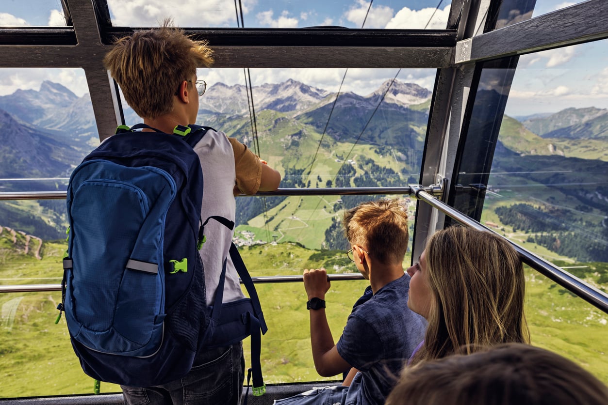 Three children ride in a glass cable car, looking out at a scenic mountain landscape with green valleys and peaks under a blue sky—one of the region’s most popular tourist attractions.