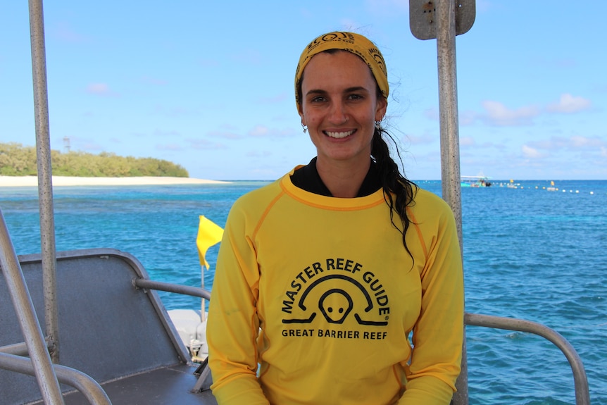A woman in a yellow rash shirt smiles while sitting on a boat with the ocean behind her