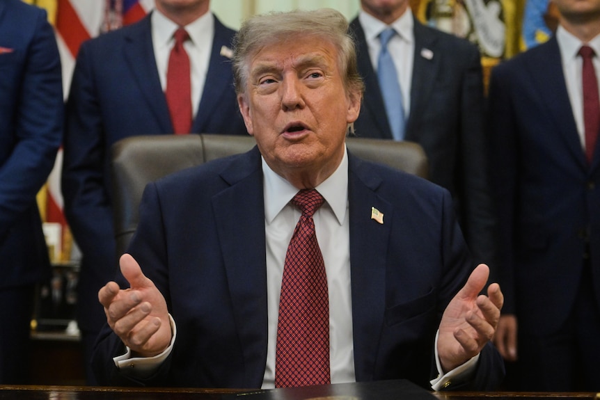 Donald Trump speaking, seated, wearing a blue suit, red tie and American flag pin.