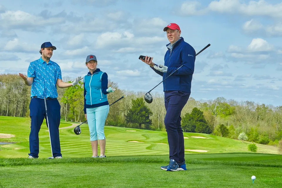 Two golfers looking at another player on the tee waiting for them to hurry up and play, with their arms outstretched in frustration