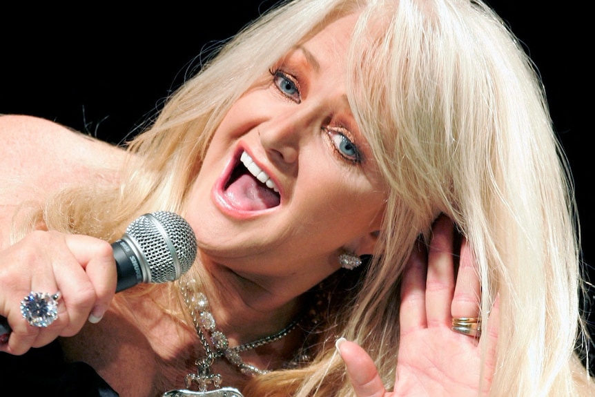 a blonde woman tilts her head while singing putting ahand up to her ear