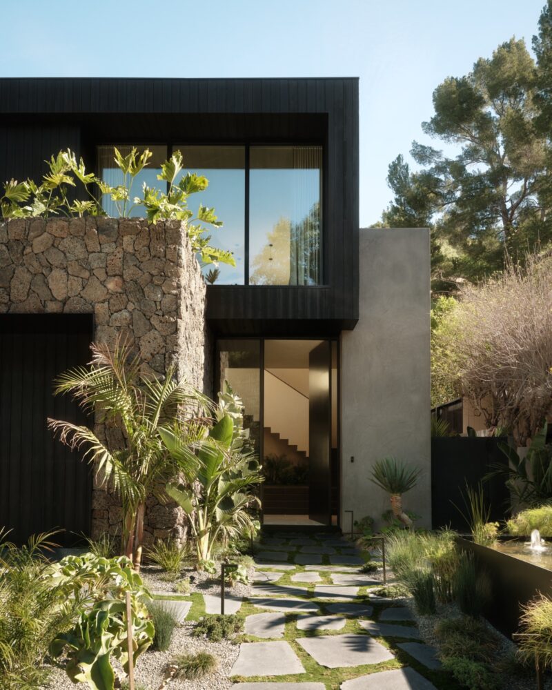 Modern two-story Obsidian House with stone and dark wood exterior, large windows, and a landscaped walkway surrounded by greenery and a small pond.
