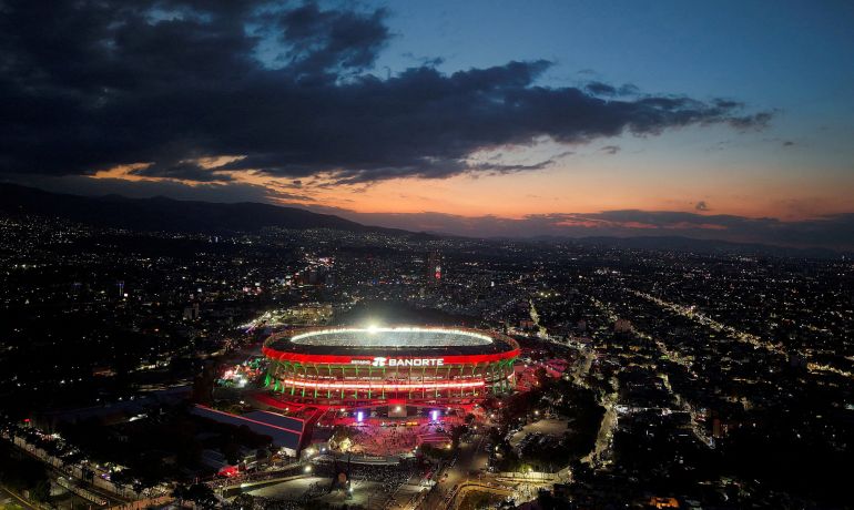 Azteca Stadium lit up at sunset with a dark city behind and clouds overhead