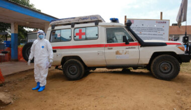 A doctor wearing personal protective equipment (PPE) stands near an ambulance