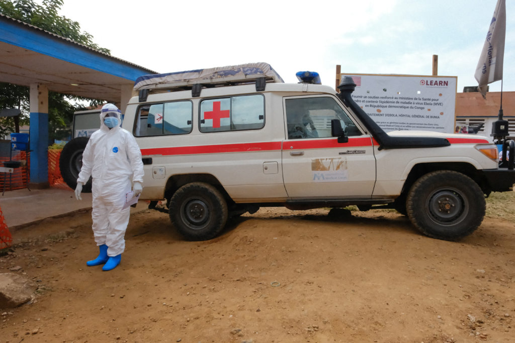 A doctor wearing personal protective equipment (PPE) stands near an ambulance