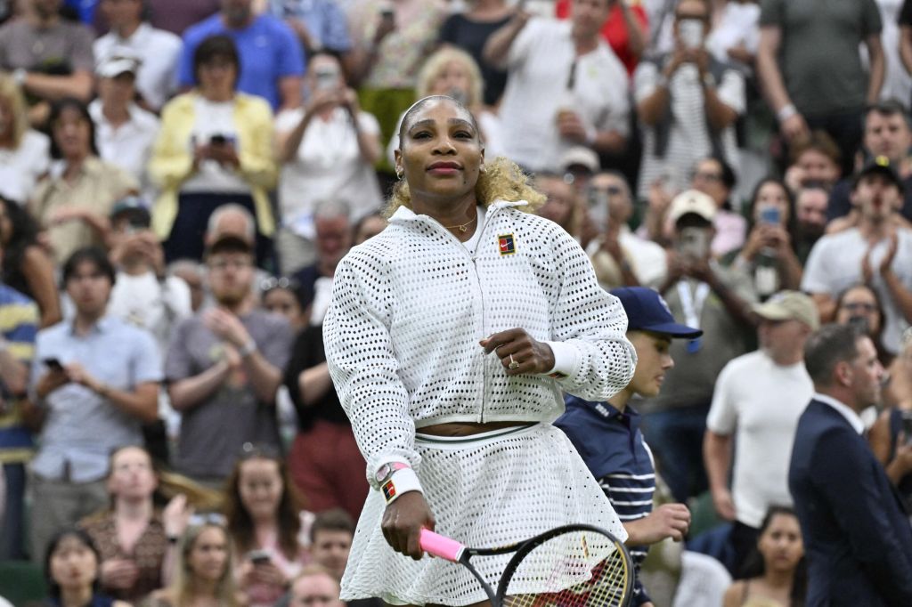 Serena Williams in a white tennis outfit with a tennis racket, walking on court, with spectators in the background.