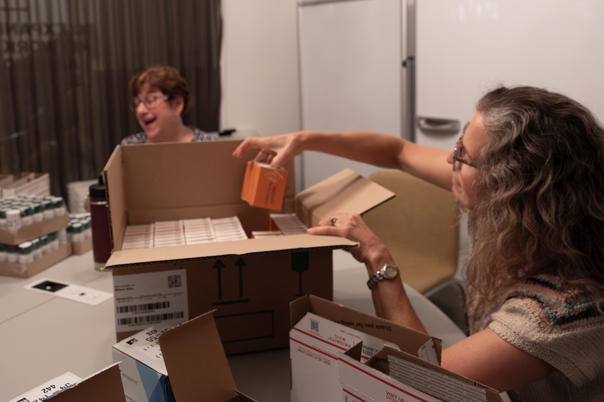 A woman seated at a table picks boxes of mifepristone from a larger cardboard box. A woman seated in the background laughs, seemingly with someone off-camera.