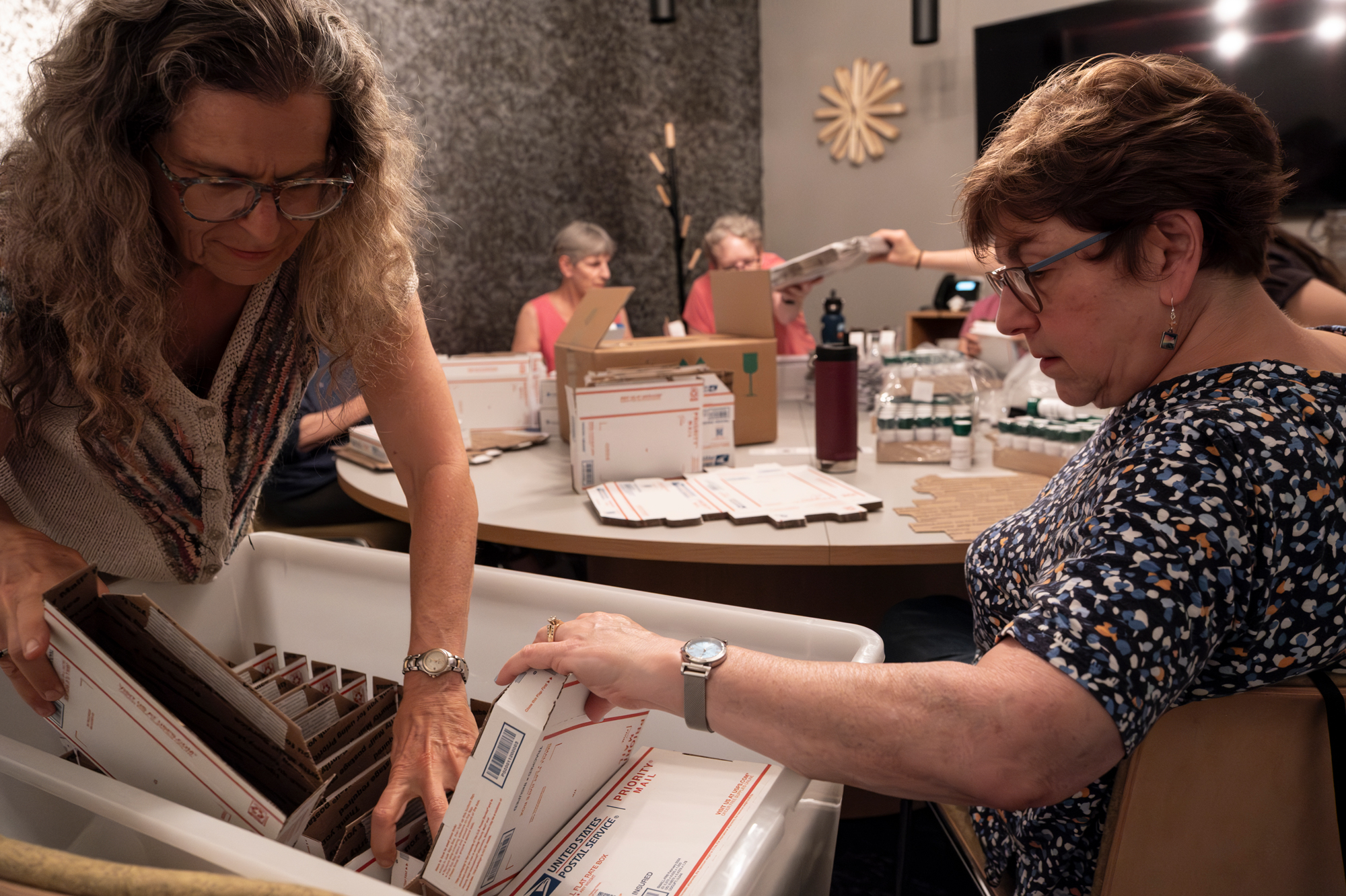 A group of women work around a table filled with supplies. Two women in focus in the foreground are handling cardboard USPS boxes in a white plastic bin.