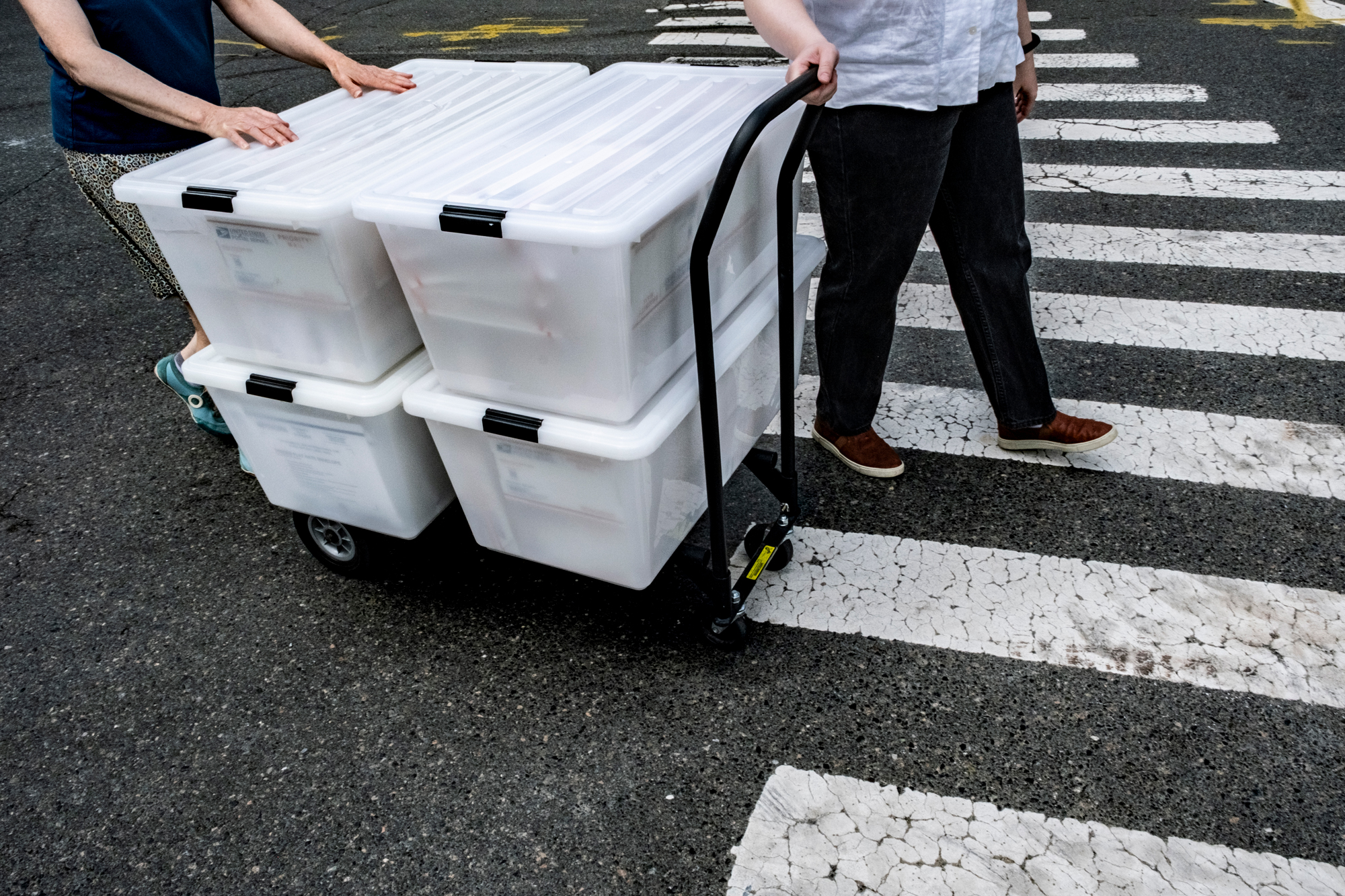 Two people, seen from the waist down, walk across a street with a cart holding four plastic bins full of USPS boxes. A person in front pulls the cart handle as a person in back helps hold the bins steady.