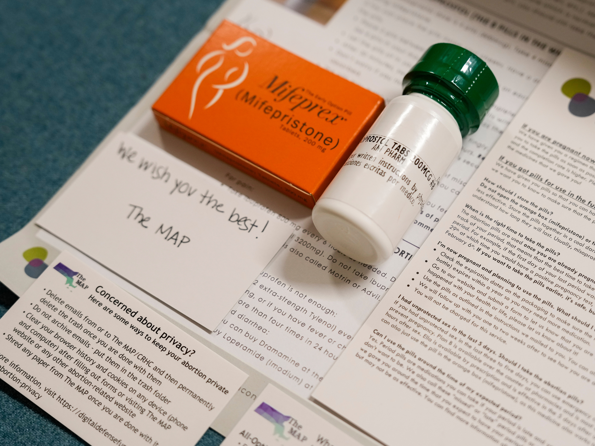 An orange box of mifepristone sits beside a white bottle with a green top. Below them are printed information sheets and a handwritten note card that reads, "We wish you the best! The MAP."