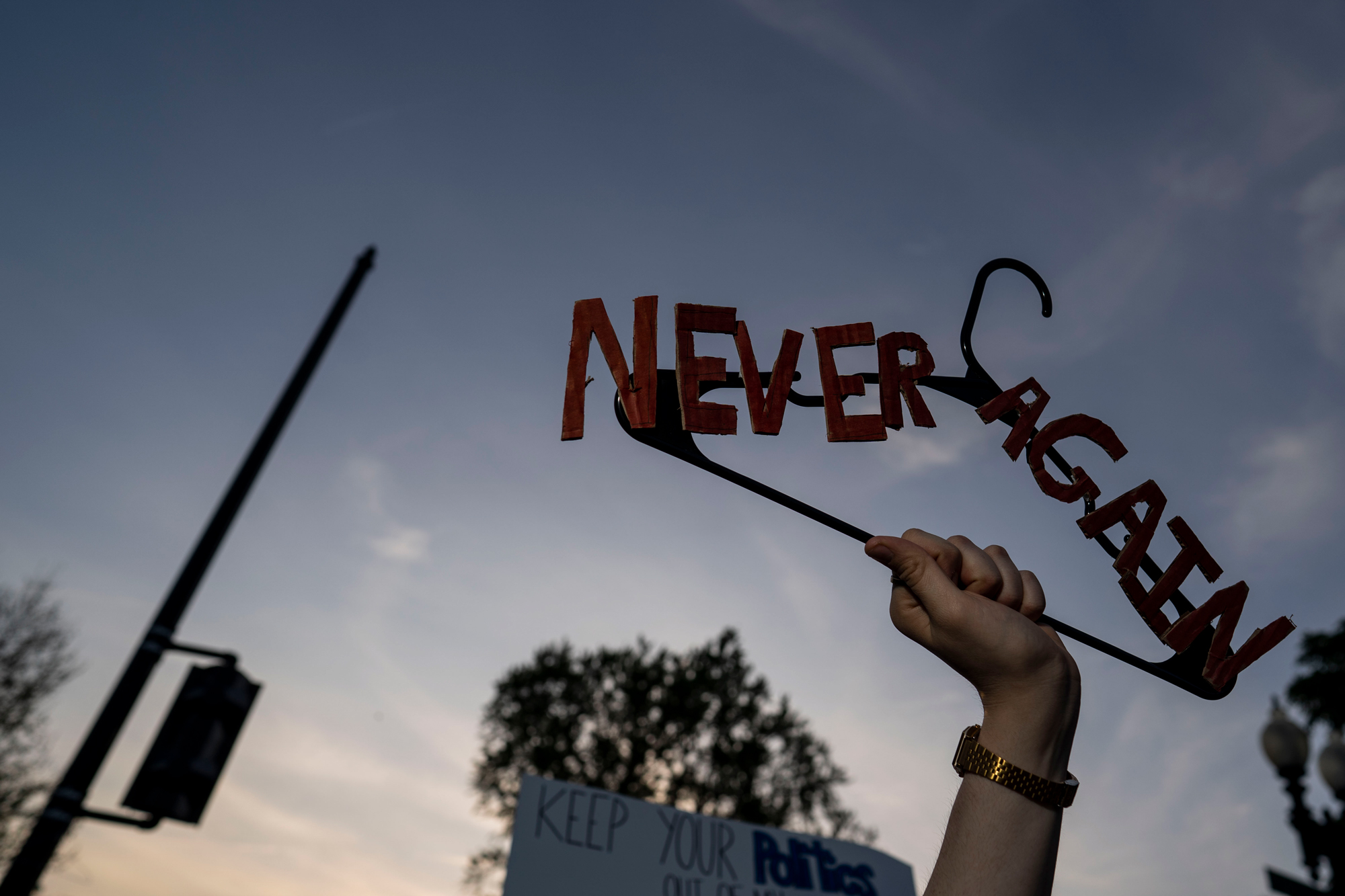 A fists raises a clothes hanger to the sky at dusk. Attached to the hanger are the cutout letters that read, "Never again."