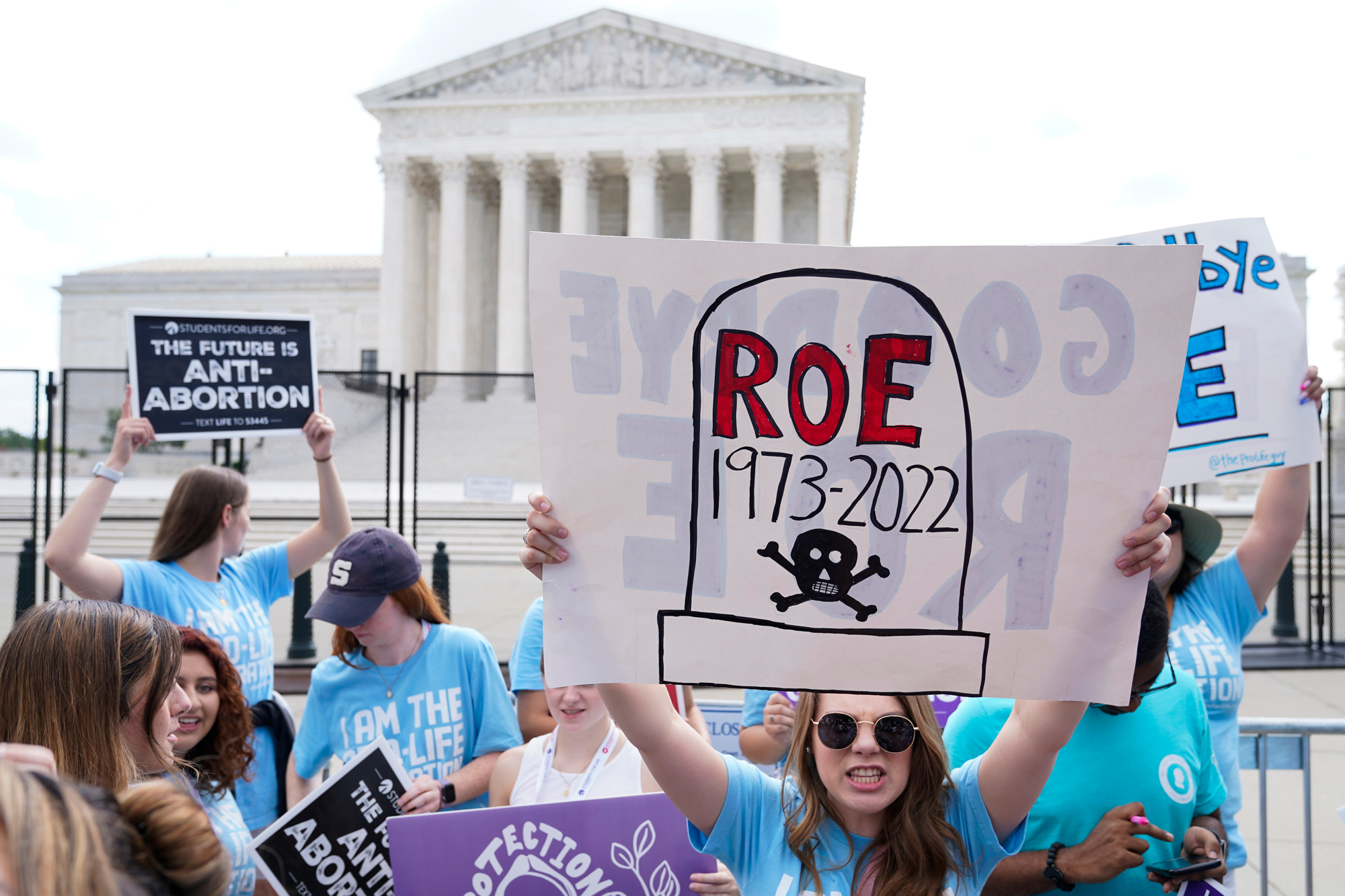 People standing in front of the Supreme Court hold signs, the most prominent reading "Roe 1973-2022" on a hand-drawn tombstone with a skull and crossbones.