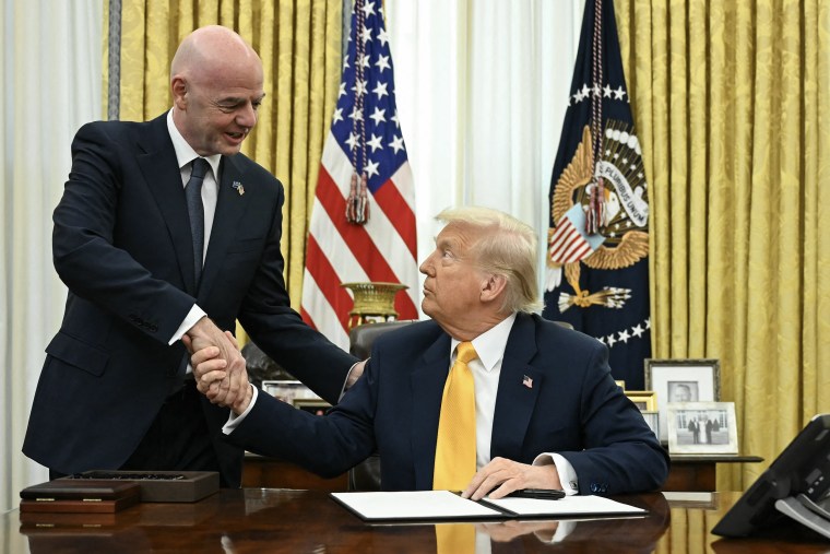 Donald Trump, seated at his Oval Office desk, shaking the hand of Gianni Infantino who stands next to him.
