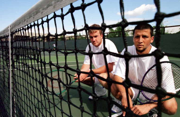 A seen through a tennis net, Ryan Wolters, left, and Paul Goldstein squat on the tennis court while holding their rackets