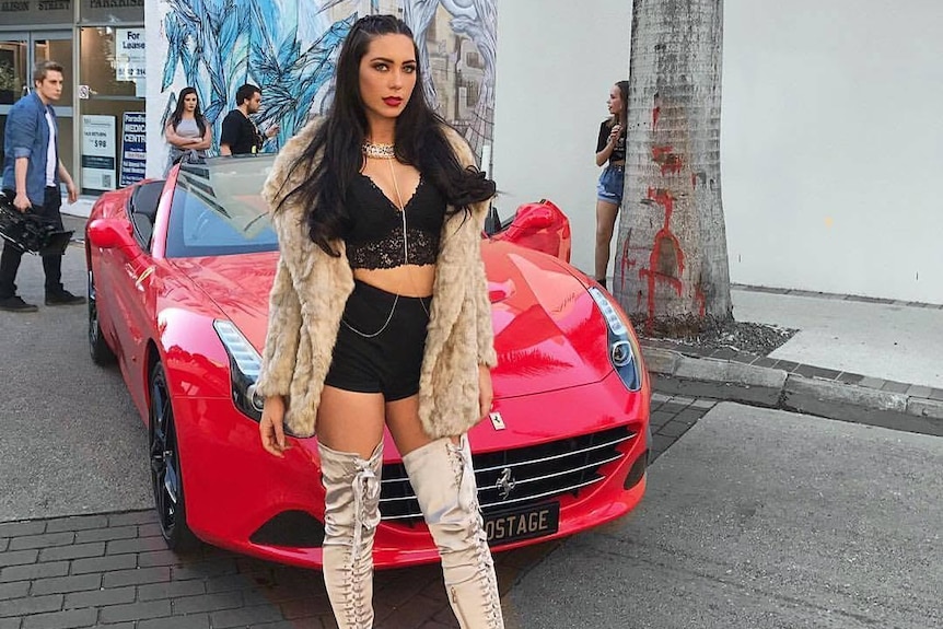 A brunette woman wearing a black crop top and skirt with a furry jacket poses in front of a red car.