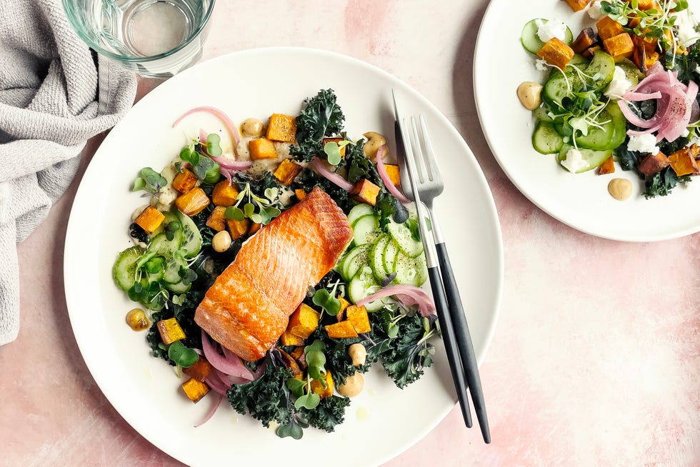 Plate of roasted salmon with fresh salad on pink background Salmon salad with kale, cucumber, sweet potatoes, and microgreens on a white plate, next to cutlery and a glass of water.