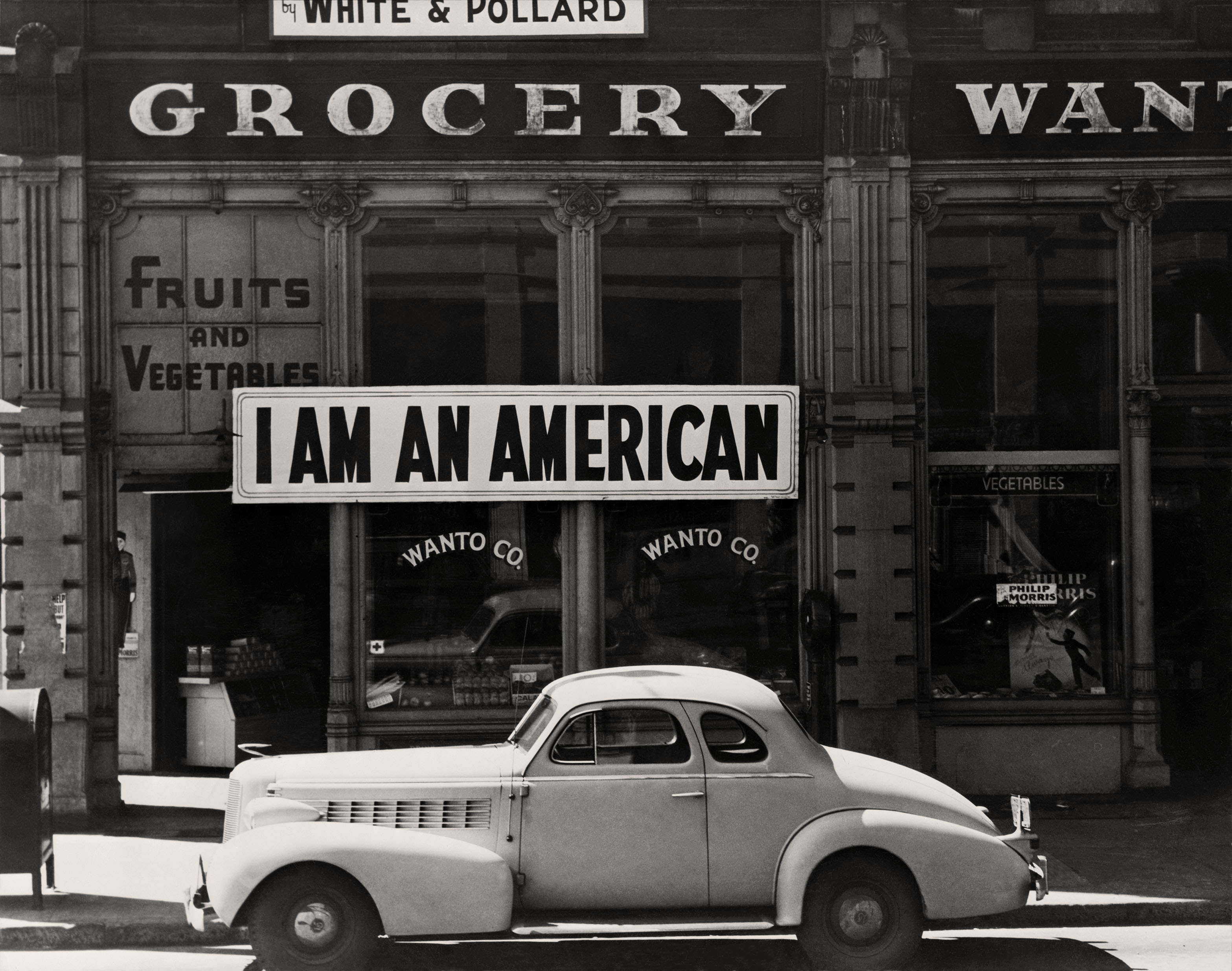 A large sign reading "I am an American" hangs in the window of a shuttered Oakland grocery store the day after Pearl Harbor, as a 1940s car sits in the foreground.