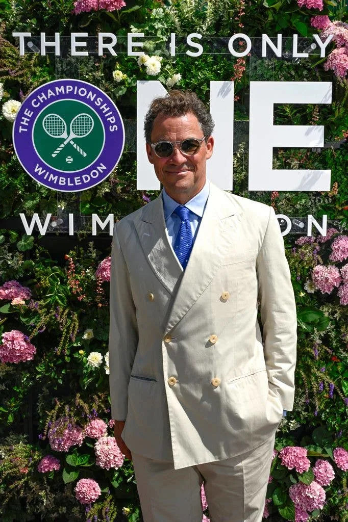 Dominic West stands in front of a floral backdrop, wearing a light suit and blue tie.