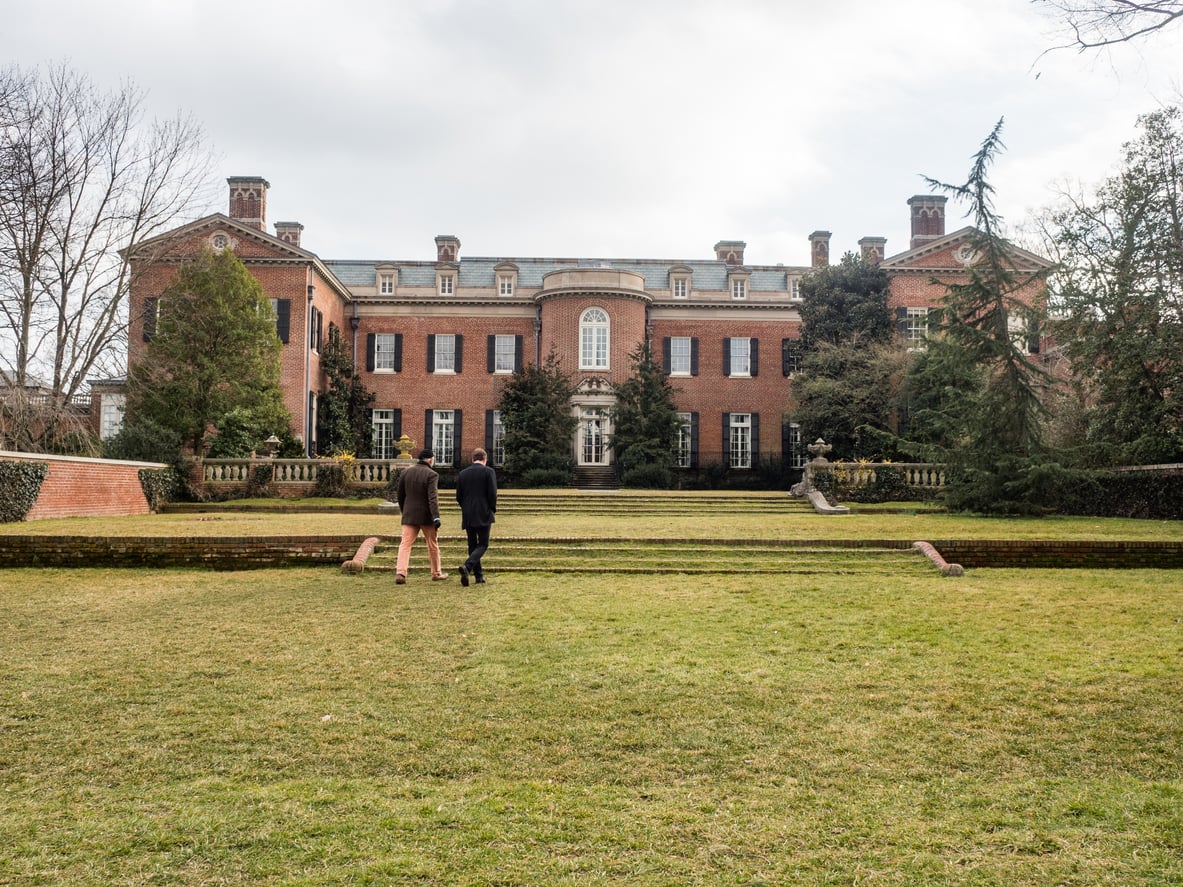 Two people walk toward a large, historic brick mansion—one of the region’s notable tourist attractions—with tall windows, greenery, and manicured lawns under a cloudy sky.