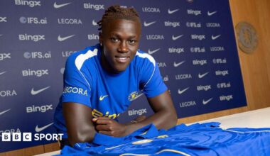Quenda resting his arms on a table with a Chelsea shirt on and in front of him after signing for the club