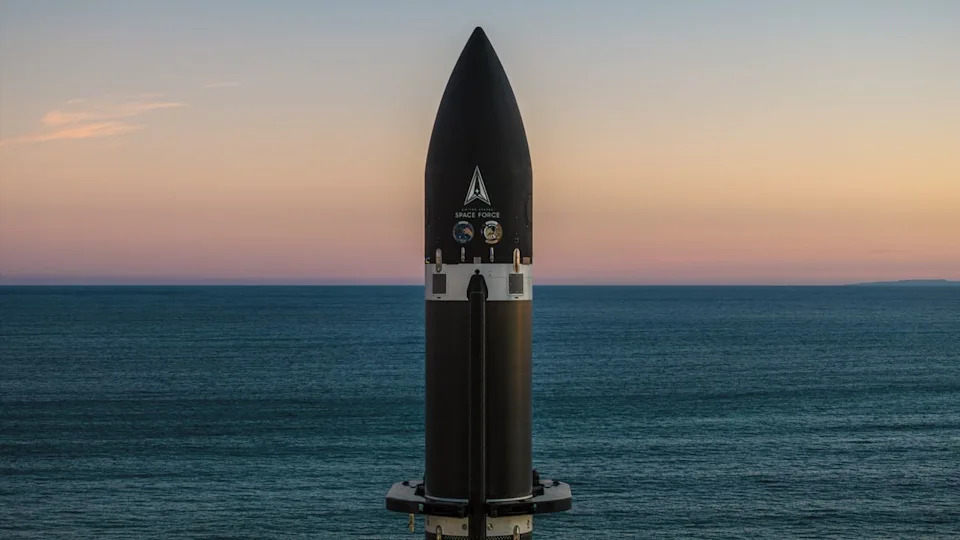 The top of a black rocket stands against a background of rich sea parted in the center by the horizon, and a cool, orange-hued sky. .