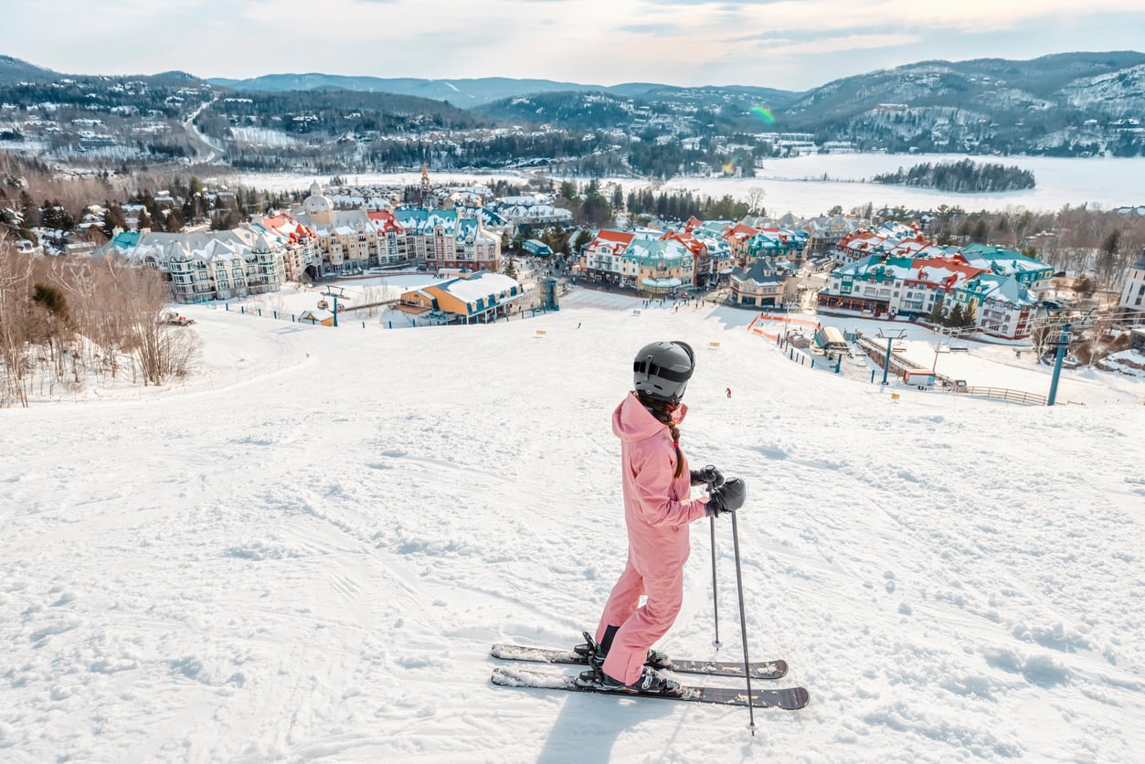 A person in a pink snowsuit and black helmet stands on skis atop a snowy slope, overlooking a colorful village renowned for its tourist attractions and forested hills under a partly cloudy sky.