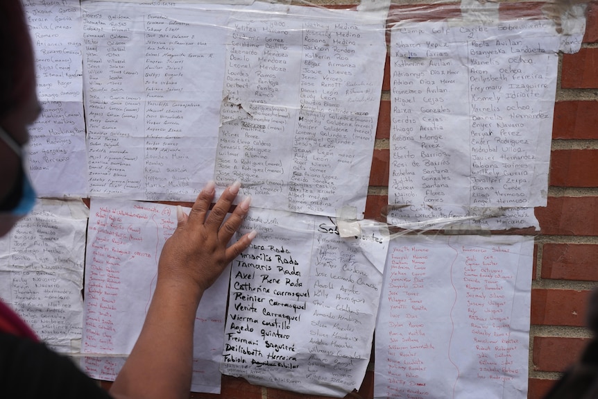 A blurred woman's silhouette, while she place her hands on white paper lists of names on a brick wall.