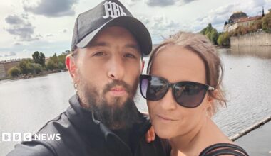 A selfie of a man and woman. On the left in the foreground, Kelvin Farmer has a brown beard and is wearing a dark coloured cap. On the right is Kayleigh, wearing dark rimmed sunglasses and her brown hair is tied back. In the background is a large body of water which could be a river or lake, and a cloudy but sunny daytime sky.