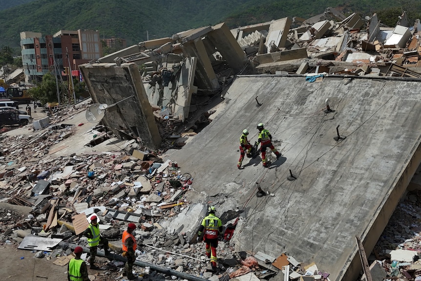 Rescuers in yellow jackets scaling a collapsed building roof amid rubble remains of buildings.