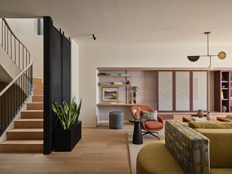 Modern living room with wooden floors, a staircase, built-in shelves, a plant, contemporary furniture, and minimalist decor in neutral and warm tones—designed as a Gillian Gillies Net Zero Ready Home for stylish and sustainable living.