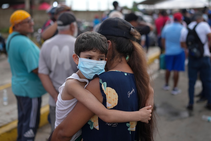 A woman in a blue top and dark cap holding a young boy wearing a blue face mask embracing her.