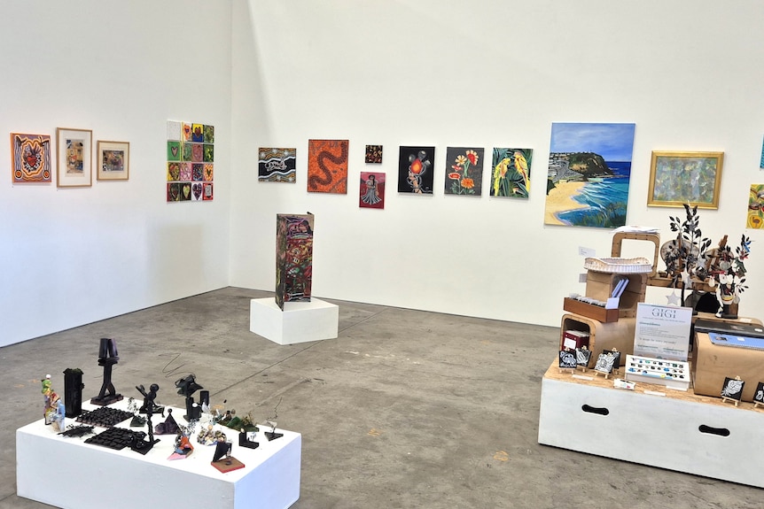An art gallery, with white walls, paintings and plinths holding objects.