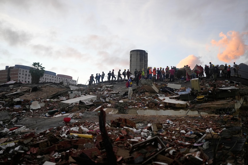 Silhouettes of a line of rescuers in hi-vis clothing and helmets standing on the horizon of a mountain of rubble.