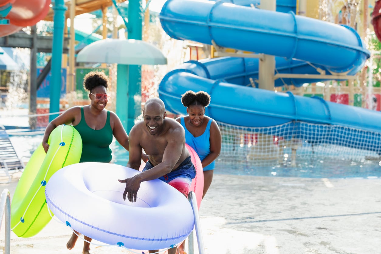 Three people, all smiling, hold colorful inner tubes as they enter a popular tourist attraction—a water park area with large blue slides and splashing water in the background.