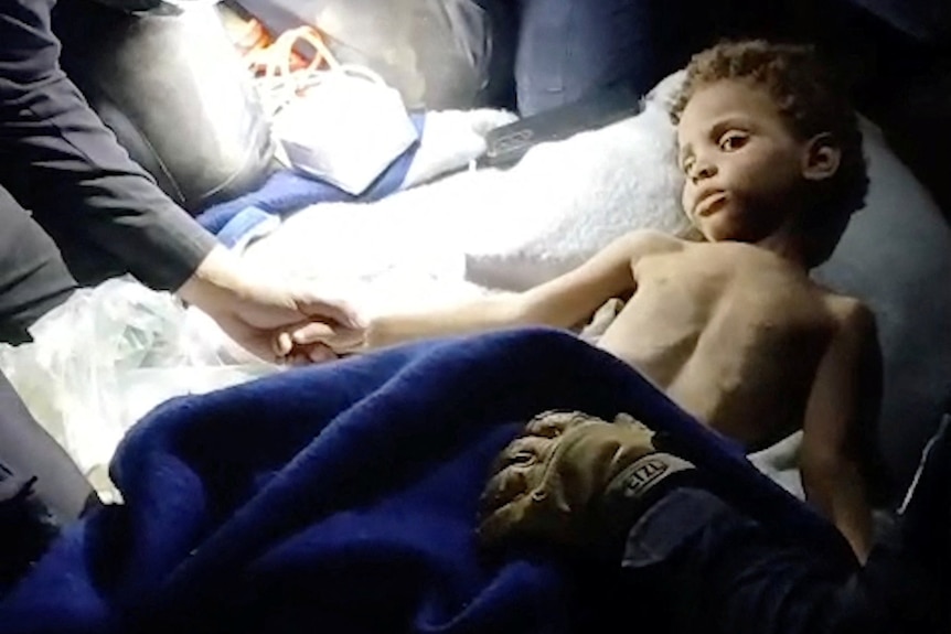 A young boy with curly brown hair appearing to be covered in dust, lying topless on a white bed, covered by a blue blanket.