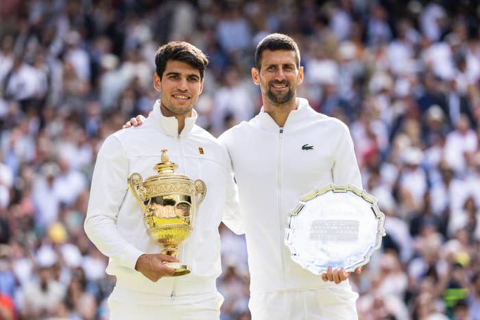 Two tennis players in white sportswear hold trophies on a tennis court, with one holding a gold cup and the other a silver plate