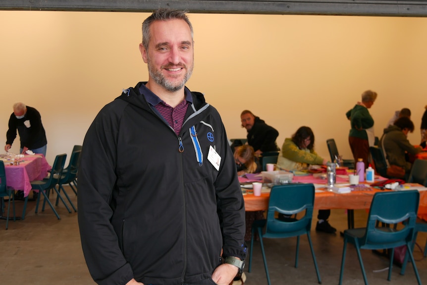 A man stands smiling while people sit at tables in the background making art.
