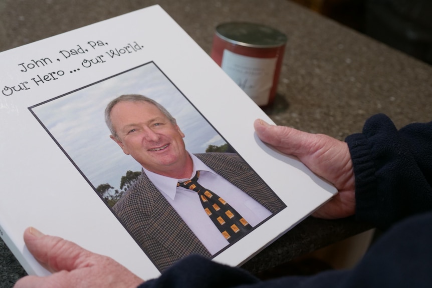 A woman holds a photo memory book with the words 'John, Dad, Pa, Our Hero ... Our World'.